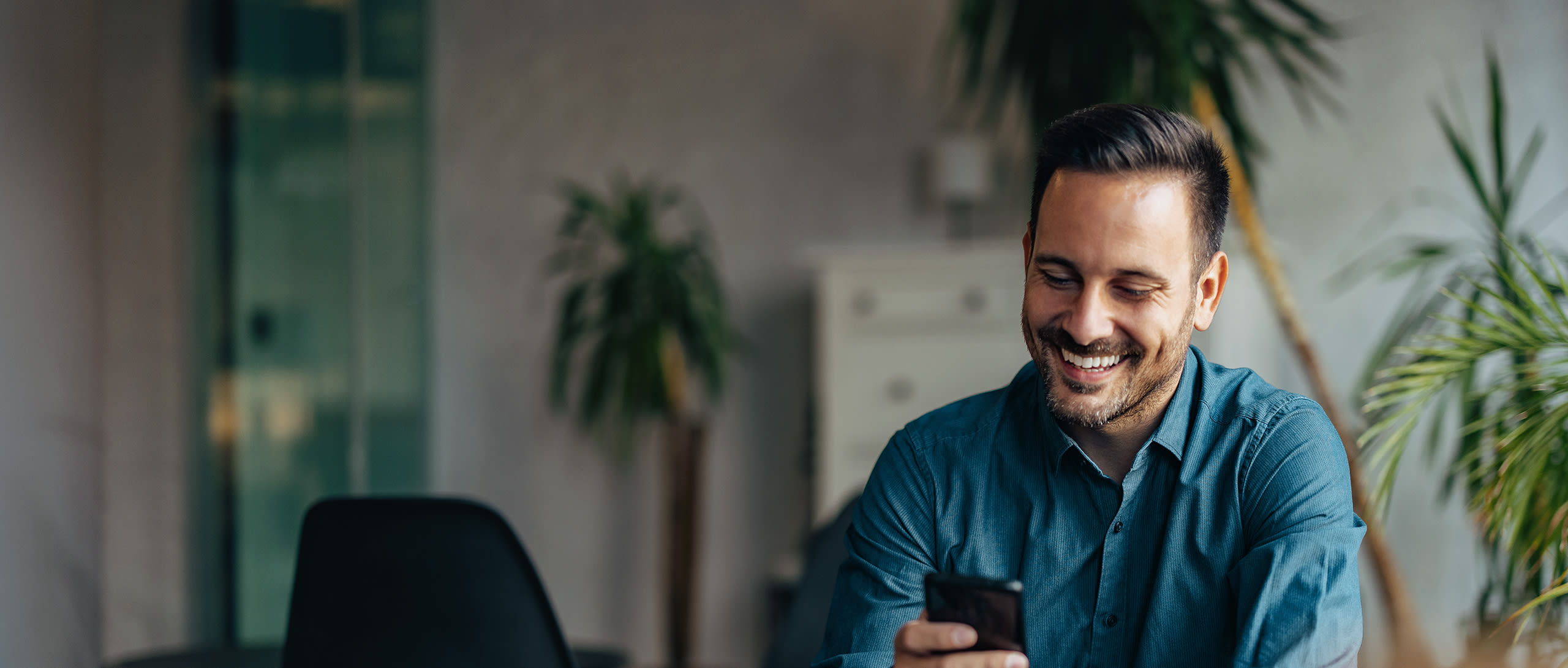 A man wearing a blue shirt smiles while looking at his cellphone