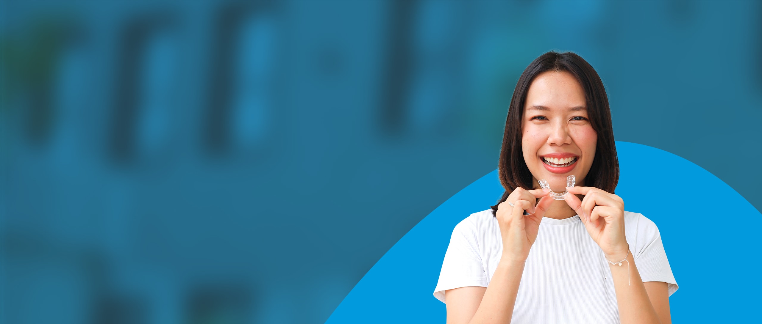 A woman is smiling and holding a clear aligners tray in front of her mouth.
