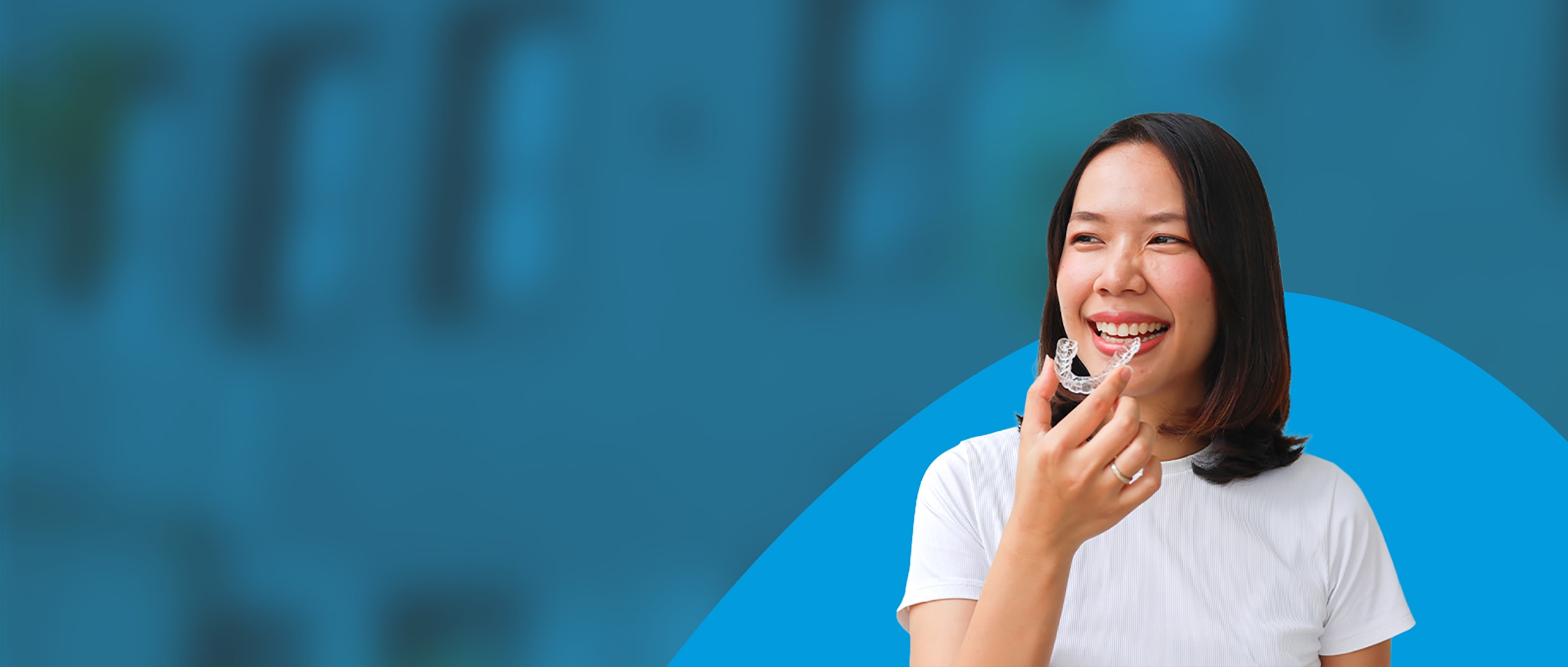 A woman is smiling and holding a clear aligners tray in front of her mouth.