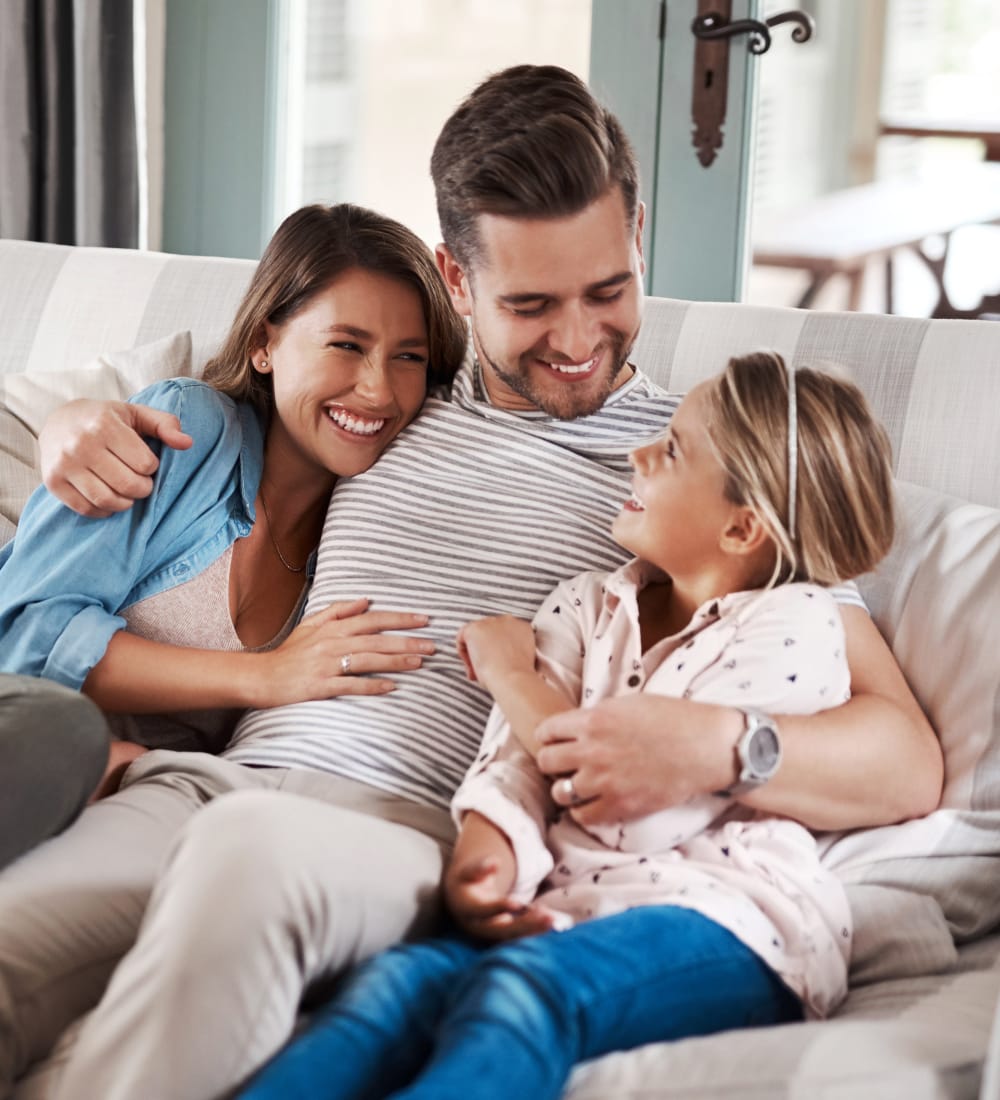 A young family, with a man, woman, and young girl hugging and smiling on a couch.