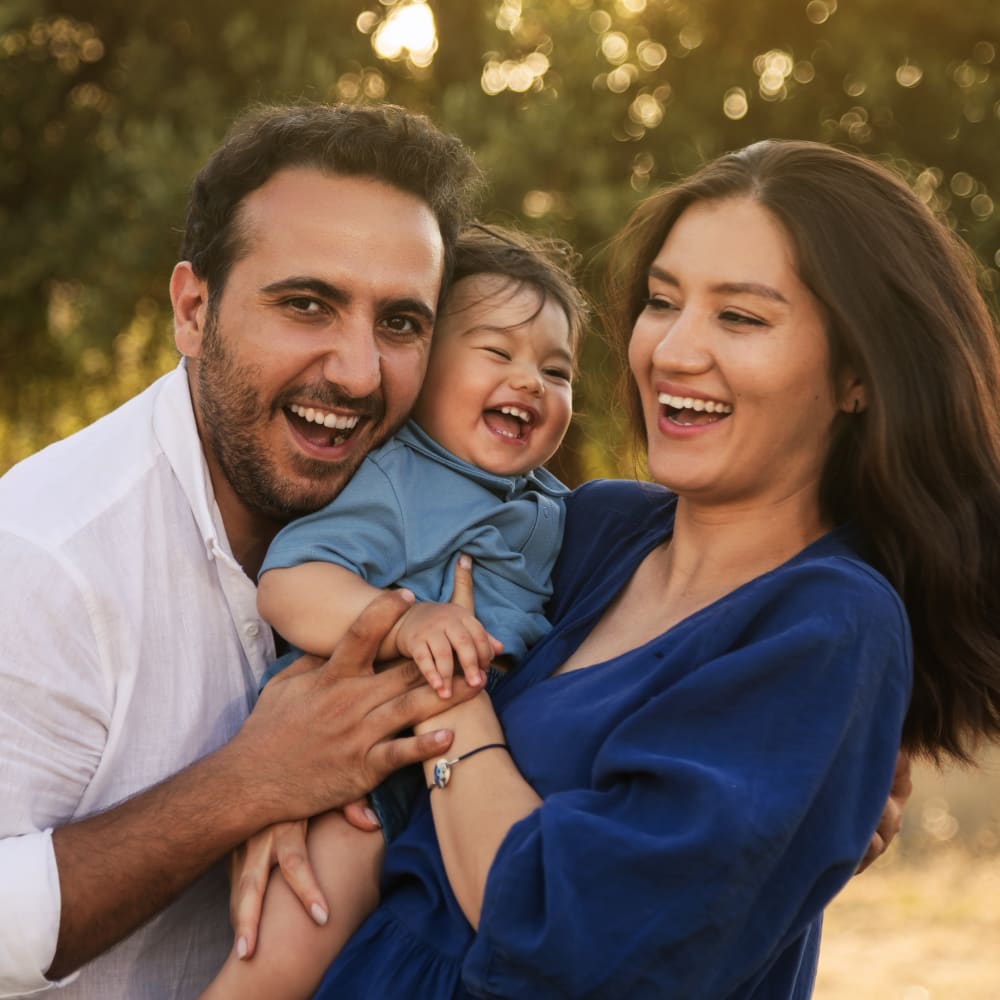 A couple are smiling and laughing. Between them, they're holding a baby that is also laughing.