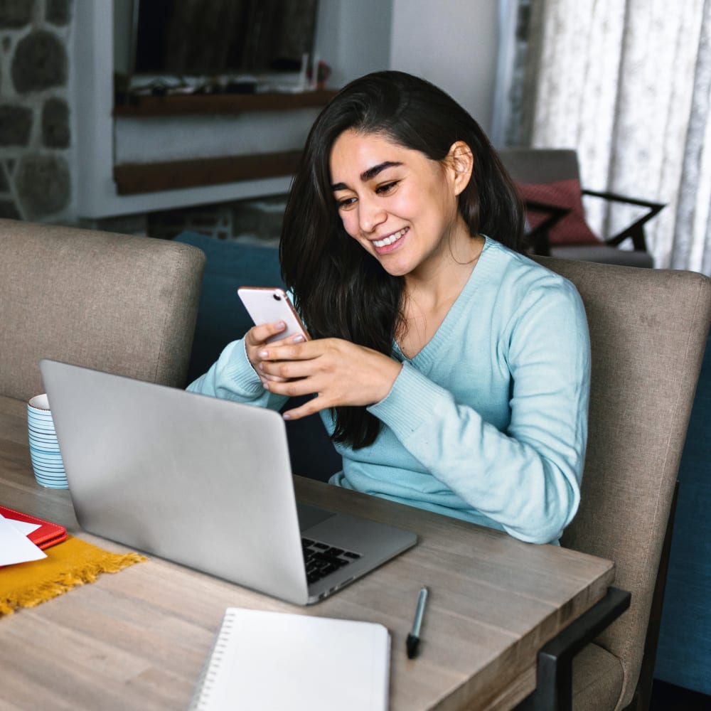 A girl is sitting at her laptop with her cell phone in her hand.