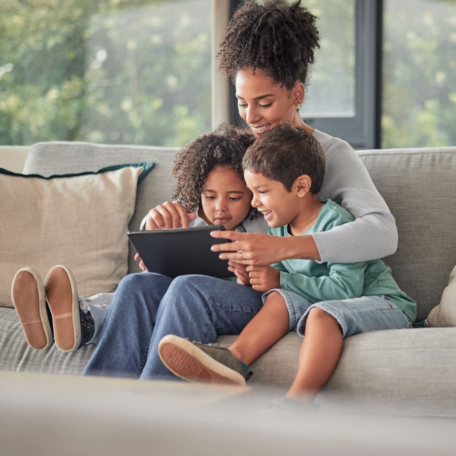 A woman is sitting on a couch looking at a tablet with two kids tucked under her arms.