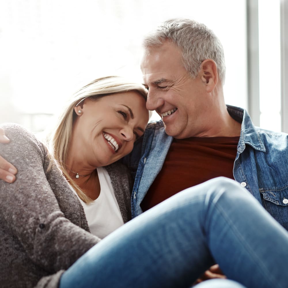 An older man and woman are smiling and embracing while sitting down. Her legs are draped across his lap.