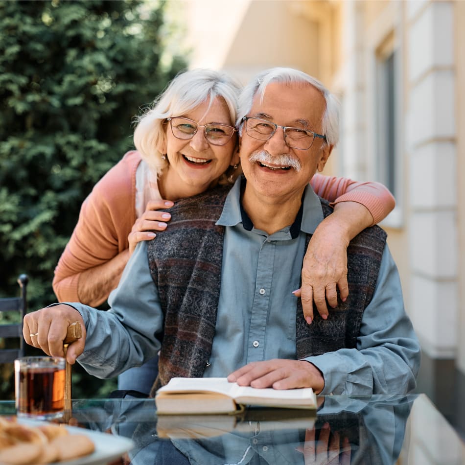 An older couple is outside. The man is sitting down smiling, while reading a book, and the woman is standing behind him with her arms around him.