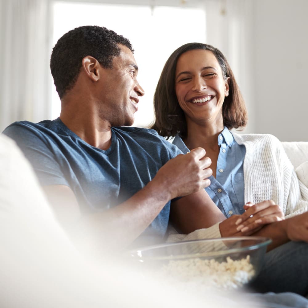 A man and a woman are sitting on a couch together smiling and eating popcorn.