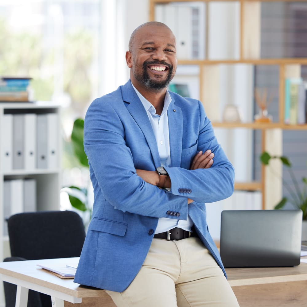 A man in a light blue blazer is standing leaned against a desk.