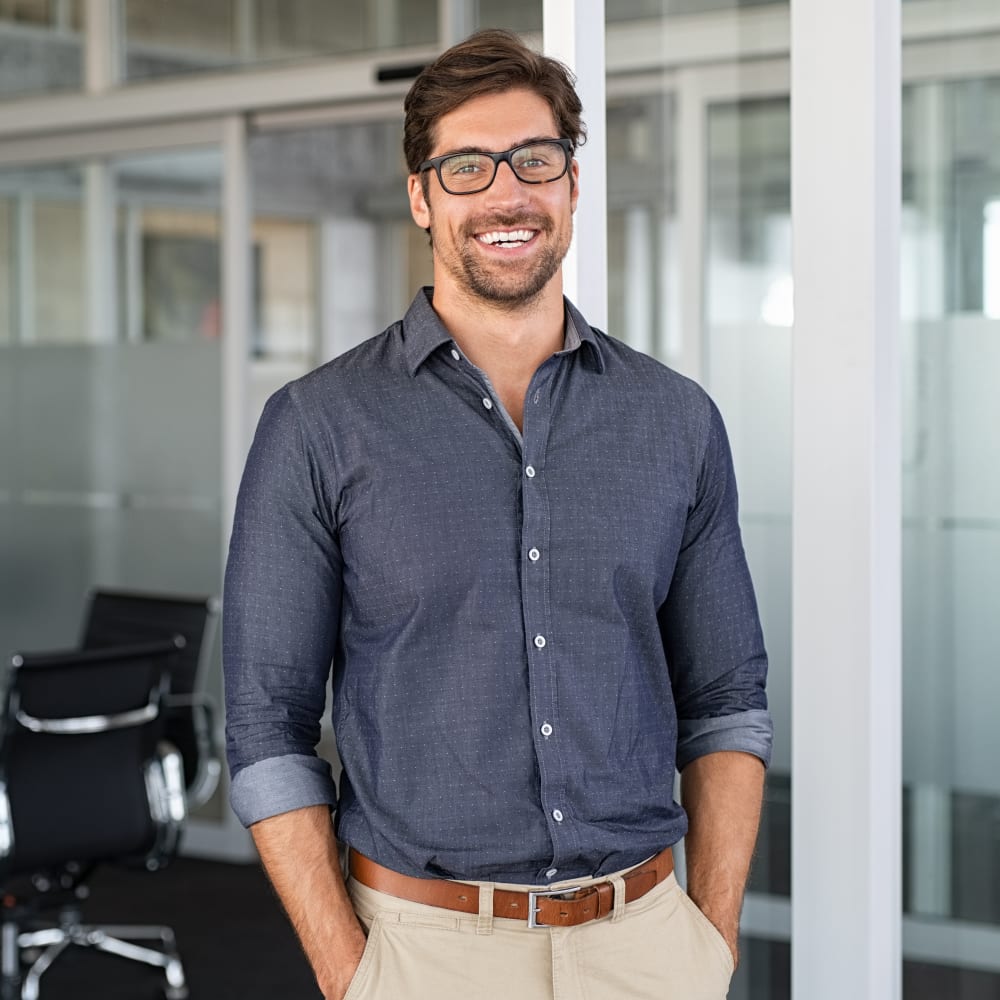 A man in a dark blue shirt has his hands in his pockets. He is smiling.
