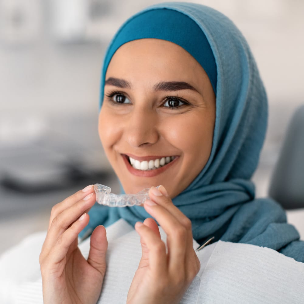 A woman wearing a head covering is smiling. She is holding an Invisalign tray up in front of her mouth.