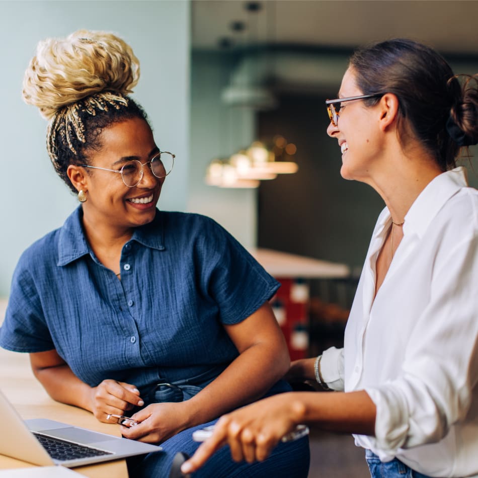 Two women are laughing together. One of them is sitting at a laptop and the other is standing beside her.