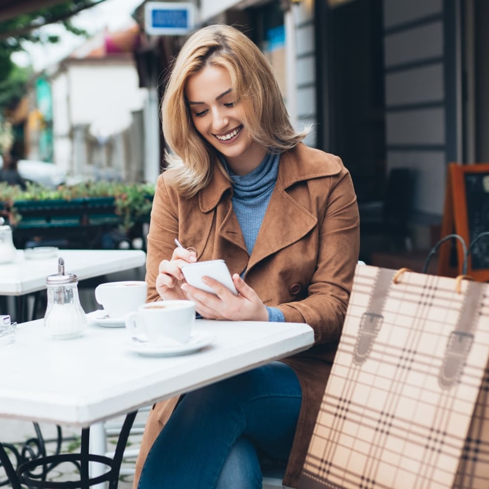 A woman is smiling and looking down at her phone. She is sitting at a table outside.