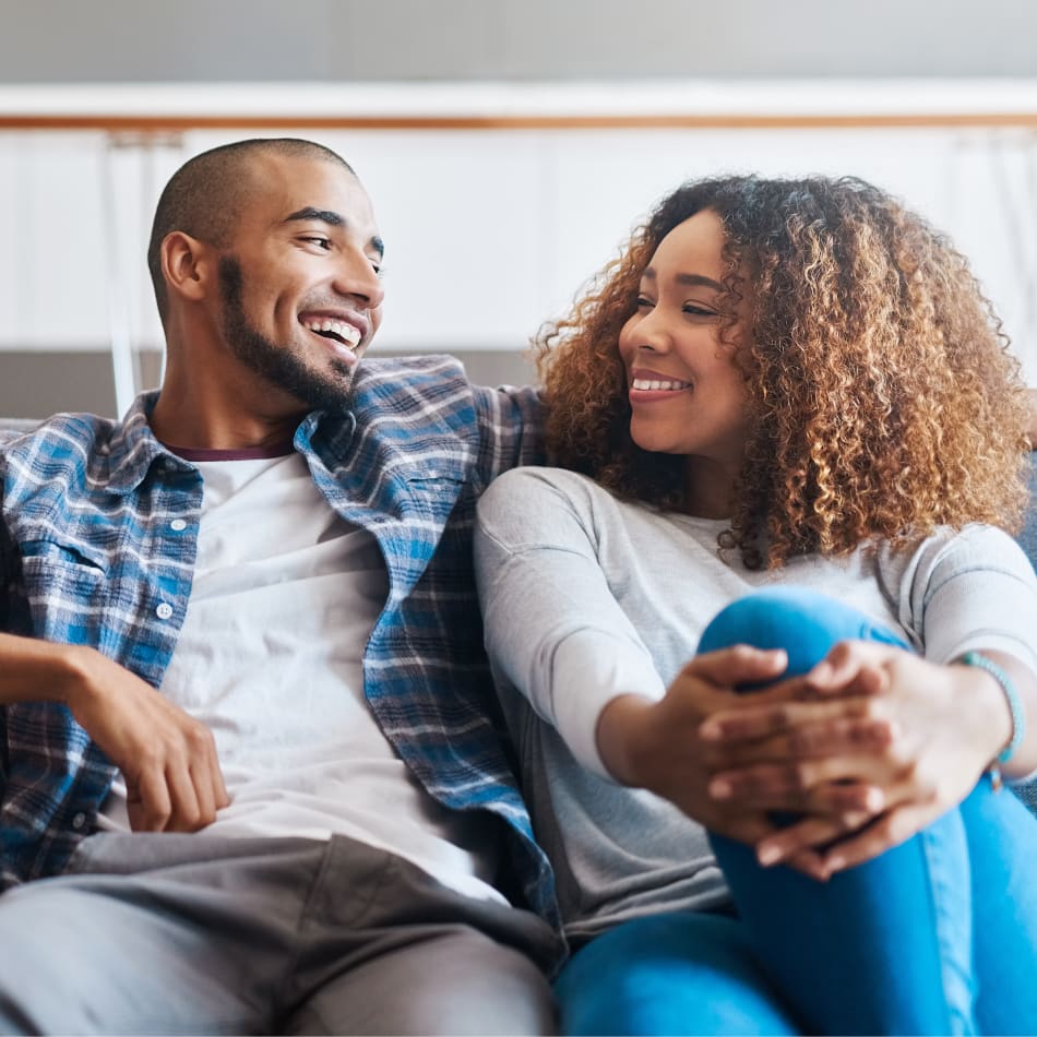 A man and a woman are sitting together on a couch. His arm is around her and she has her hands clasped over her knee.