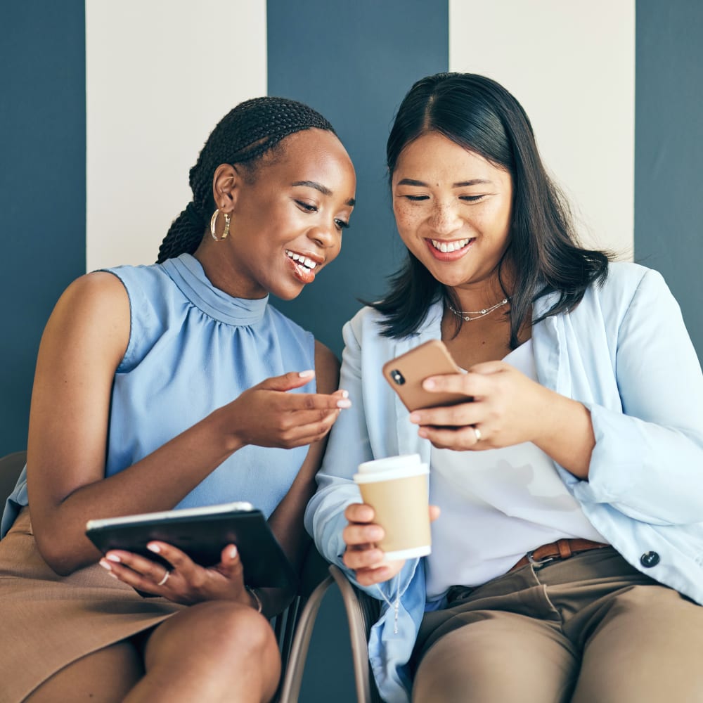 Two women are sitting together, chatting, and looking at one of the women's phones.